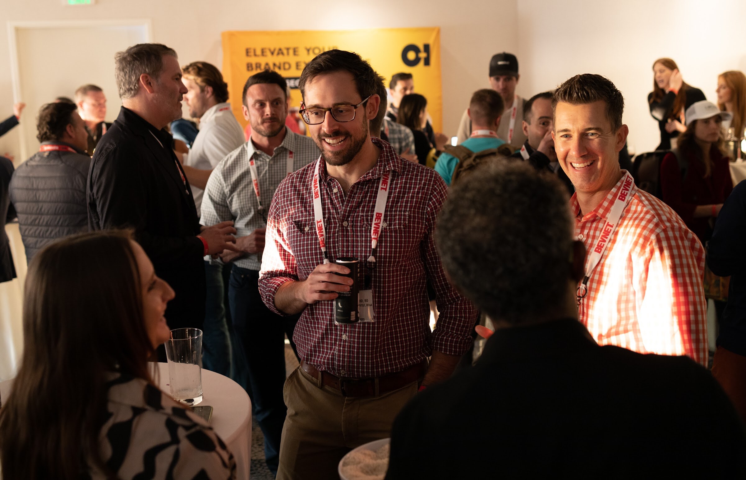 A group of attendees in plaid shirts and BevNET lanyards smile and talk in a crowded room with an O-I banner behind them.