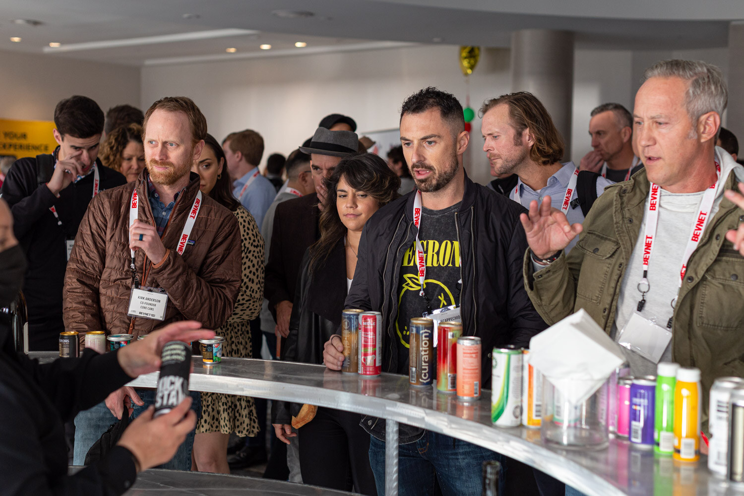 A crowd stands around a curved counter with many canned drinks, including a man in a Nirvana t-shirt and a man in a green jacket.