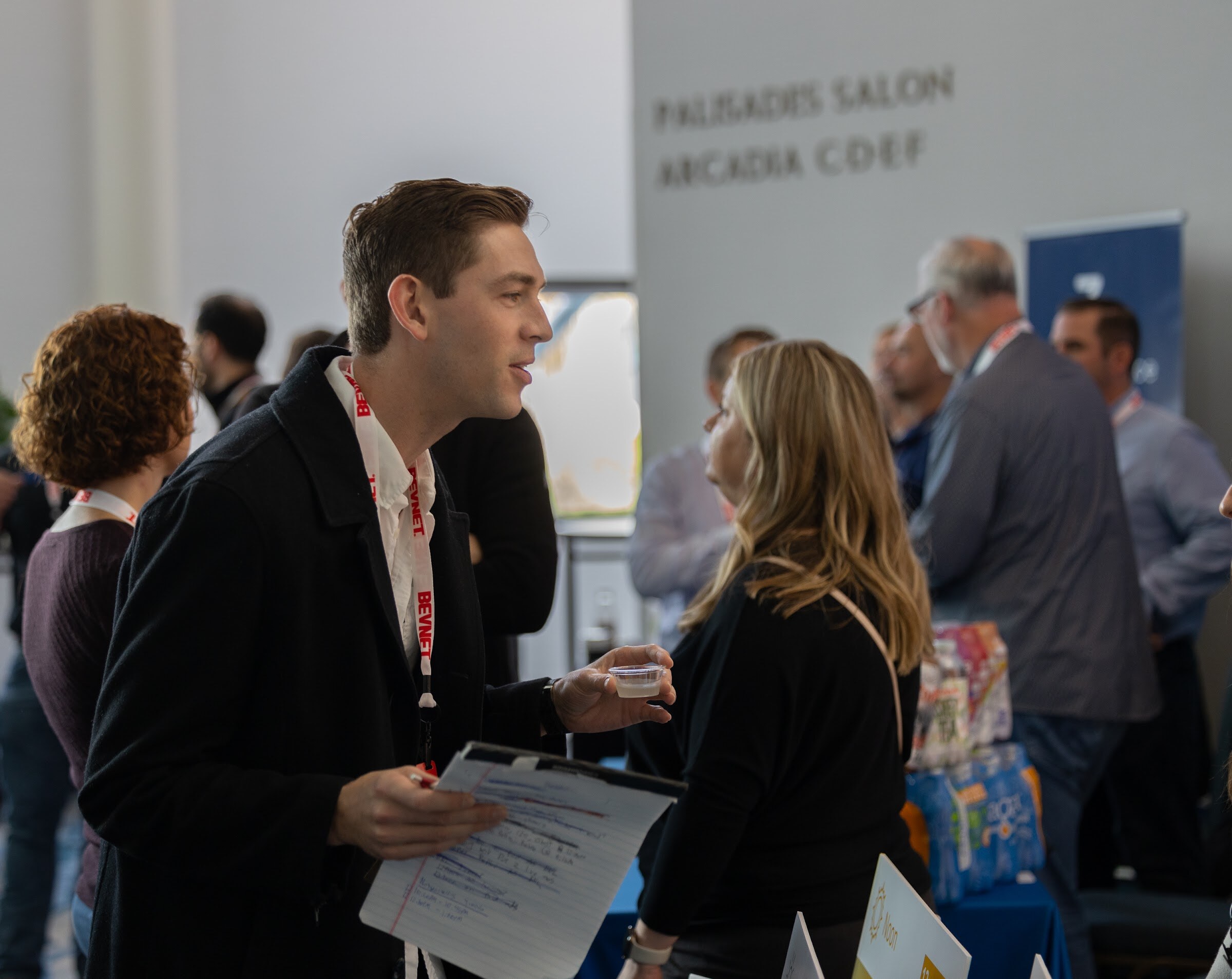 A man holding a clipboard and a sample cup talks to a woman with long blond hair in a crowded hotel hallway with a sign reading Palisades Salon.
