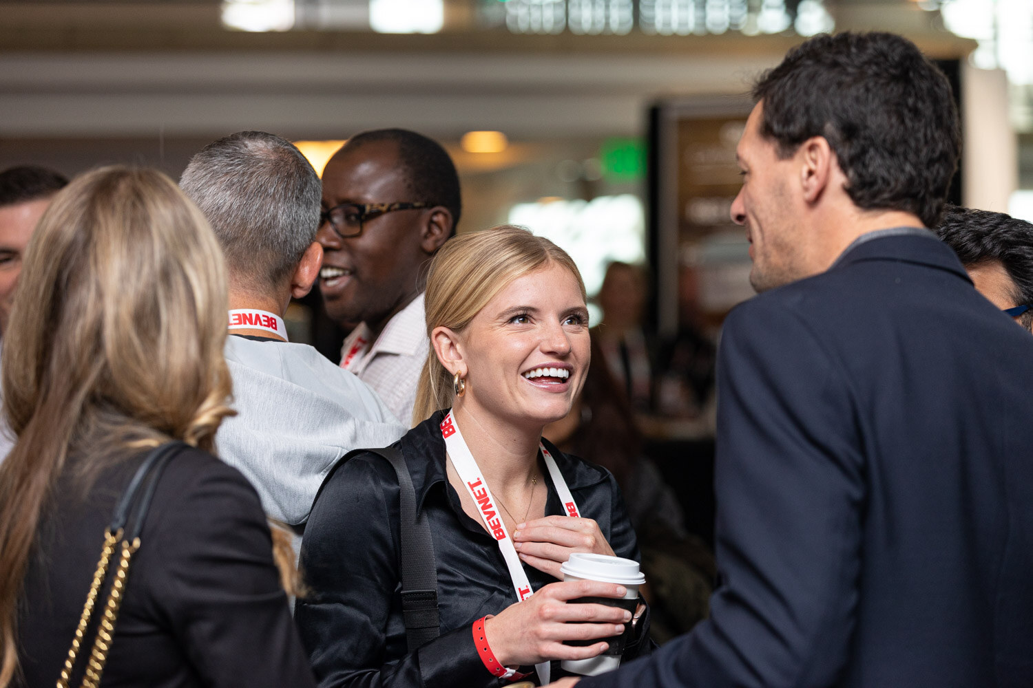 A smiling blond woman in a black blouse with a BevNET lanyard holds a coffee cup while talking with a man in a navy blazer.