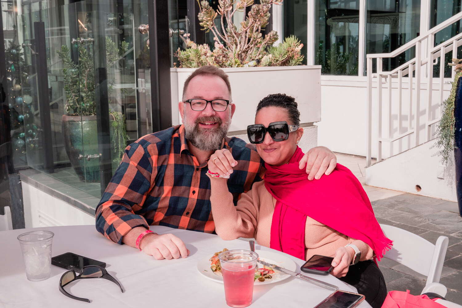 A bearded man in glasses and a plaid shirt and a woman in sunglasses and a pink scarf pose at a table with salad and a pink drink.