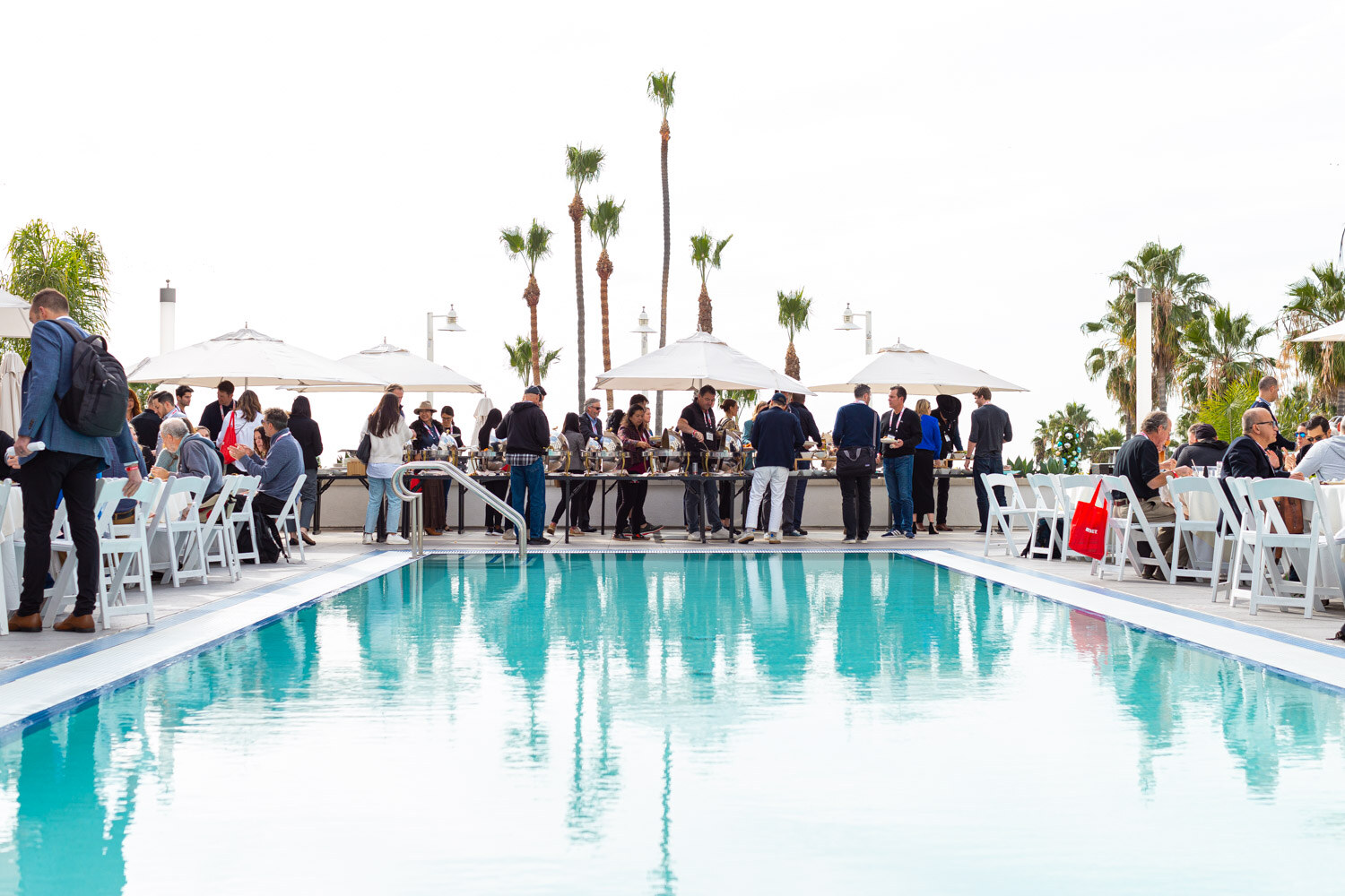 A bright blue swimming pool with palm trees and attendees gathered at a buffet on the far side under white umbrellas.
