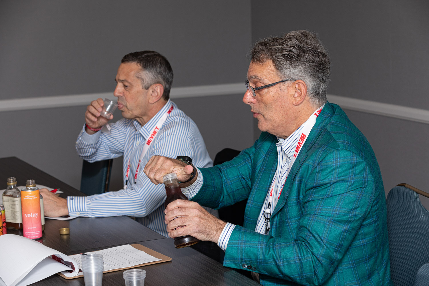 Two men in BevNET lanyards sit at a table, one in a teal plaid blazer holding a small bottle and the other sipping from a cup, with several bottles and a can on the table.