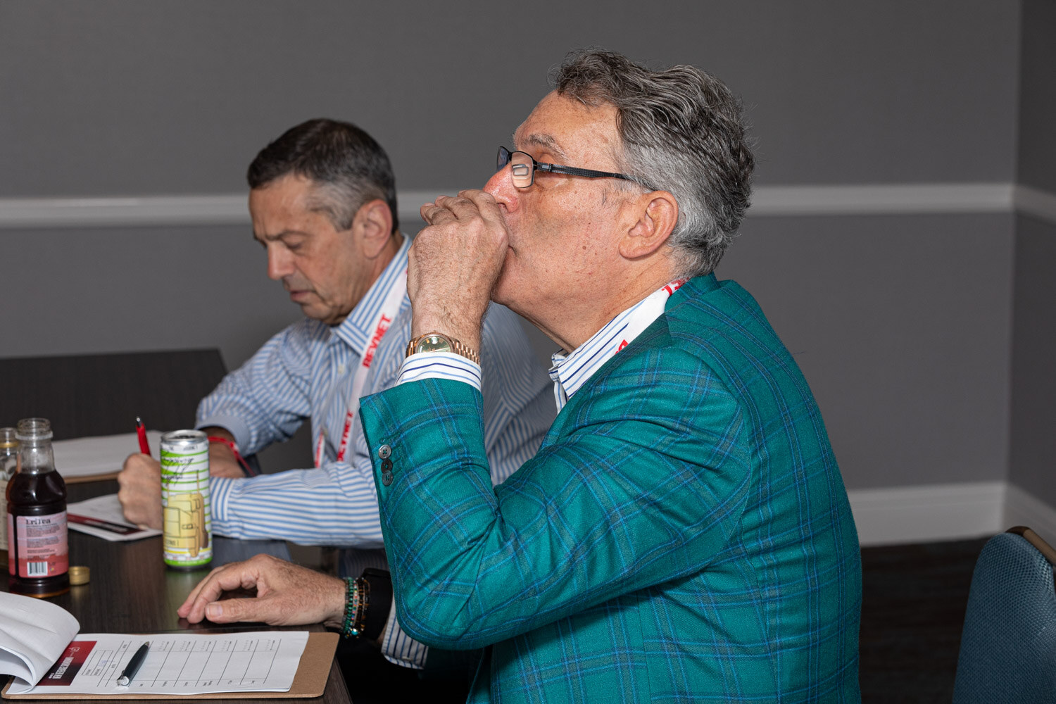 A man in glasses and a teal plaid blazer sips from a small cup at a table with scoring clipboards, bottles and a can, with a second man writing beside him.