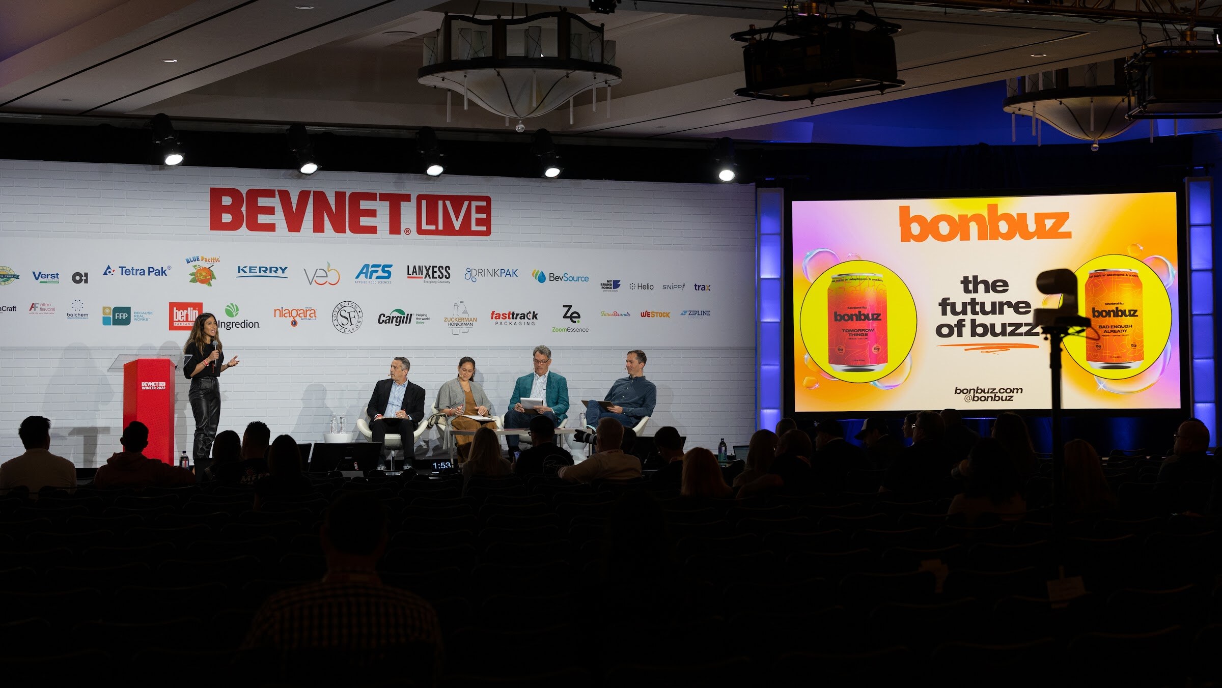 A wide shot of the main stage in a dim ballroom, with a presenter standing beside a panel of four seated judges and a bonbuz slide on the screen.