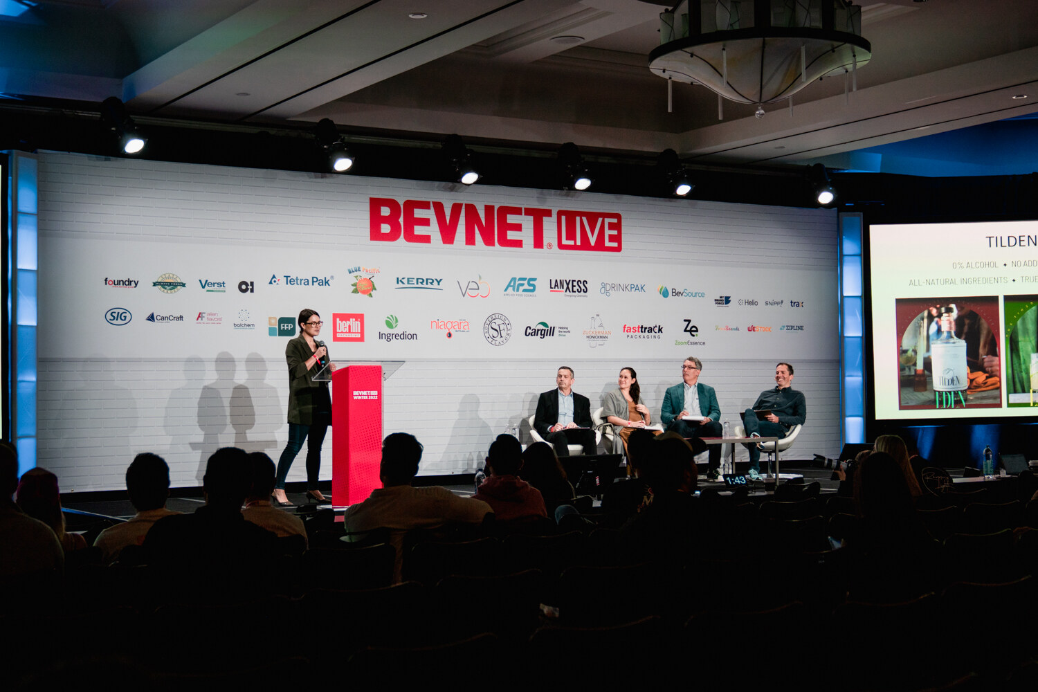 A woman stands at a red podium on the left of a dark stage while four judges sit in chairs under a BevNET Live sign.