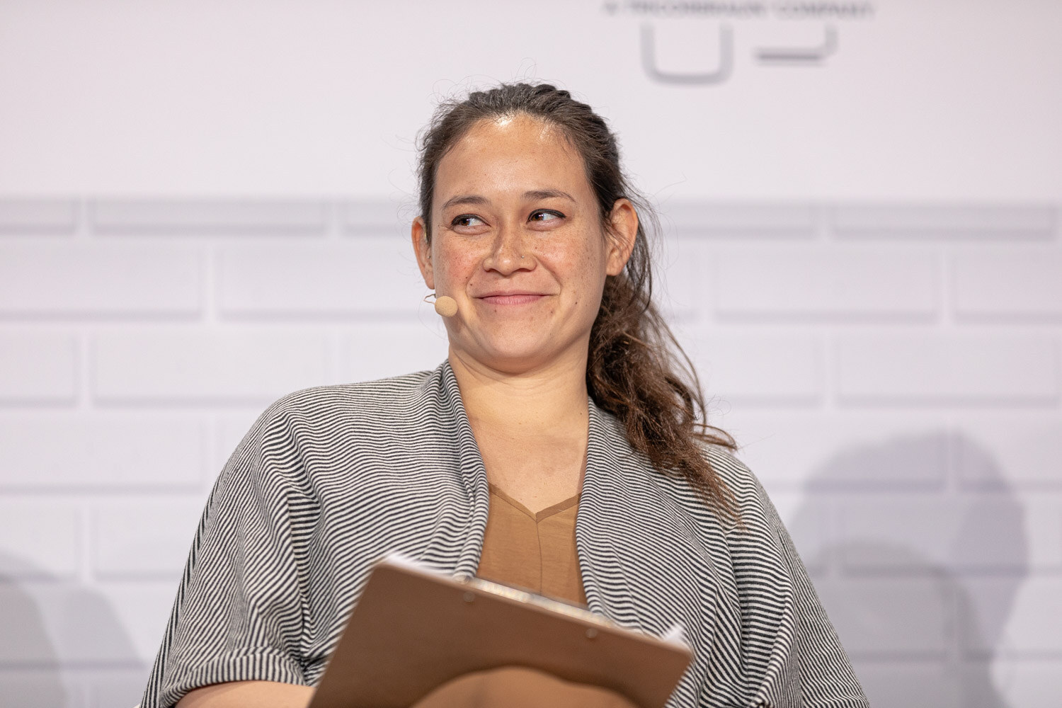 A woman in a gray striped cardigan holding a clipboard and wearing a headset microphone smiles in front of a white brick wall.