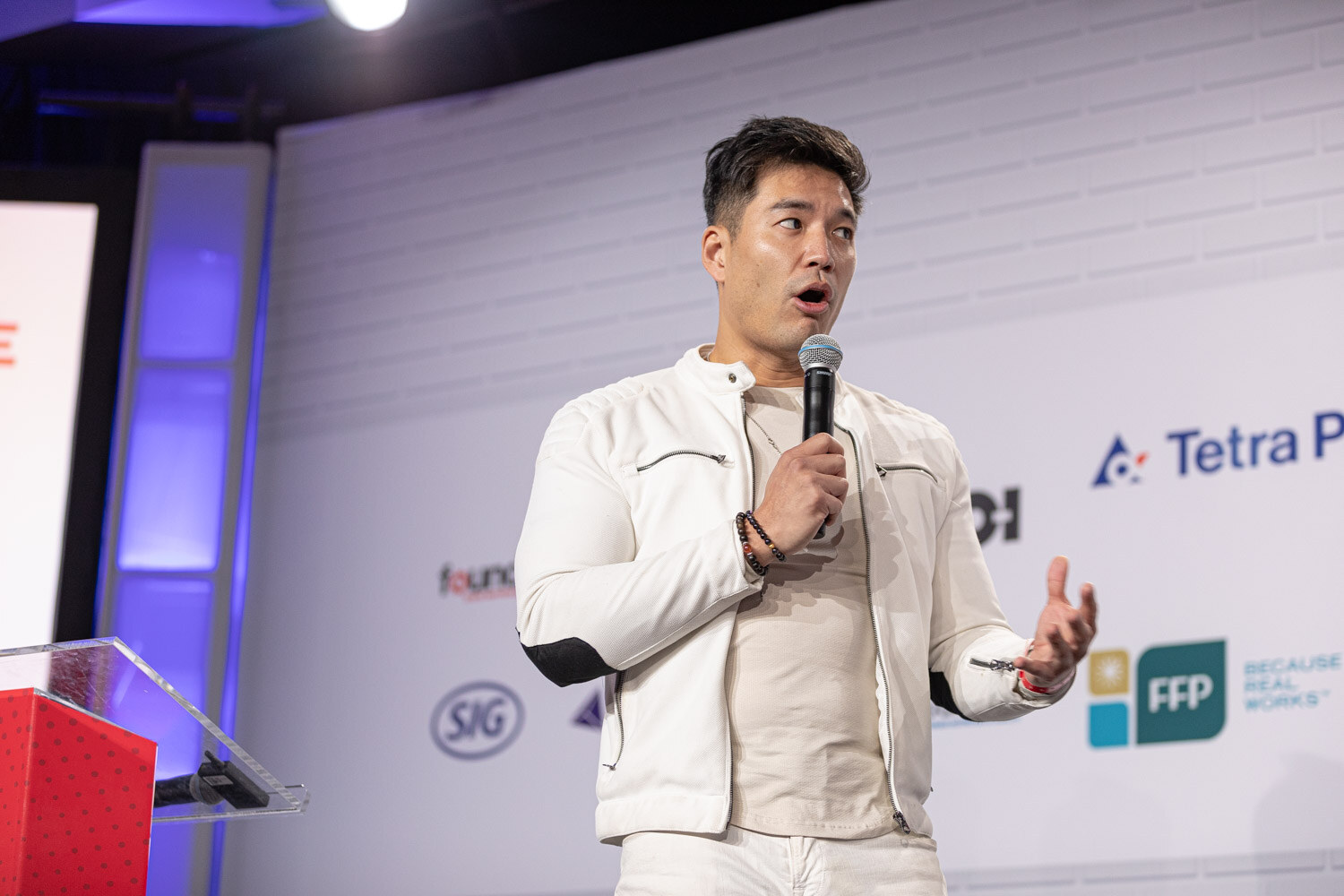 A man in a white jacket holds a microphone beside a red podium on stage in front of a sponsor wall.