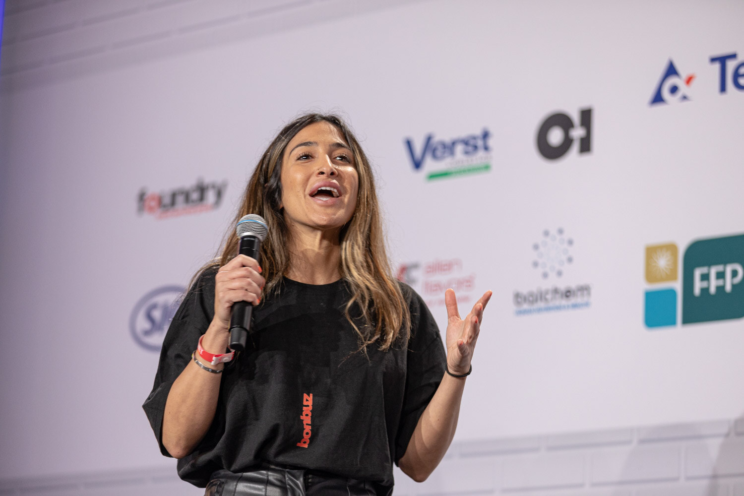 A woman in a black t-shirt with bonbuz lettering holds a microphone and gestures with an open hand on the stage.