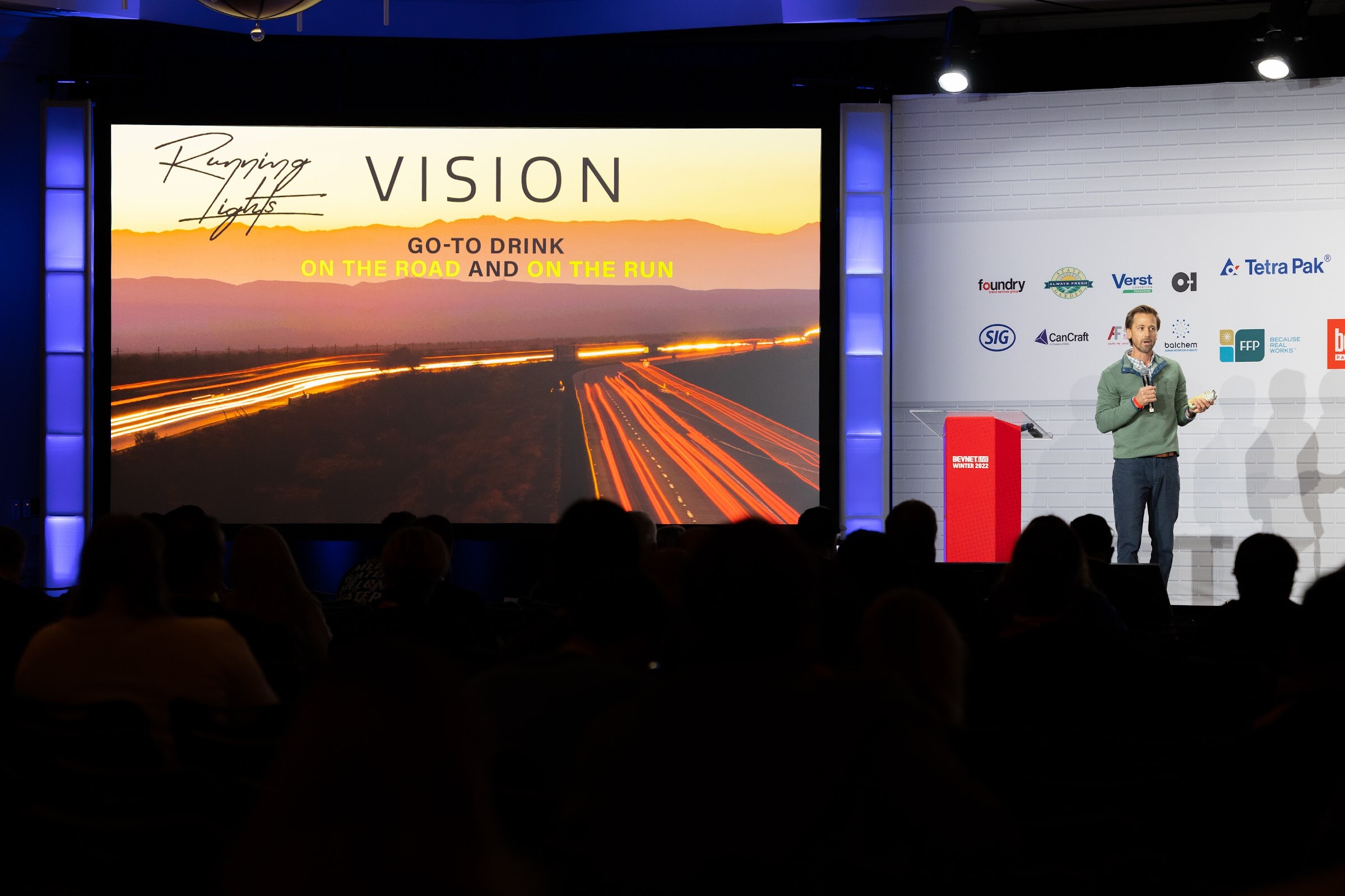 A man stands on the stage beside a podium in front of a large screen reading Running Lights Vision, Go-to drink on the road and on the run, with a darkened audience in the foreground.
