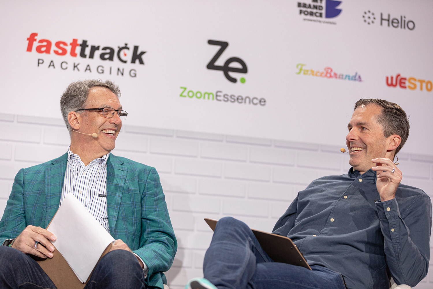 A man in a teal plaid blazer and a man in a navy shirt, both wearing headset microphones, laugh in white chairs in front of a sponsor wall.