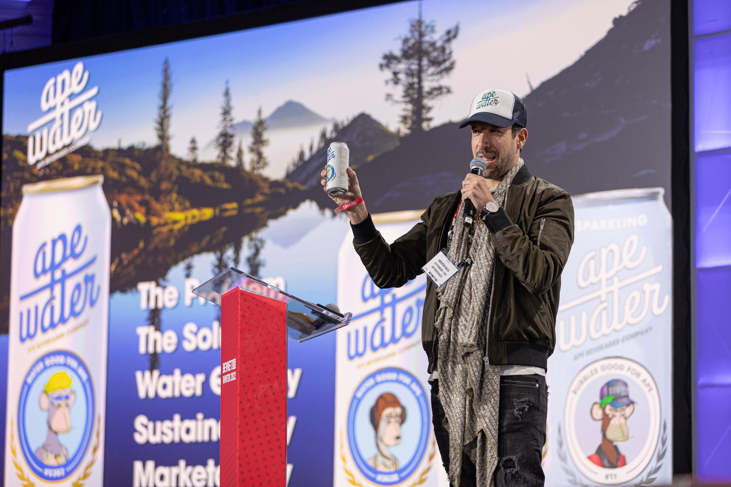 A man in a ball cap and green jacket holds a can and microphone on stage in front of a screen reading ape water, The Problem, The Solution, Water Quality, Sustainability, Marketability.