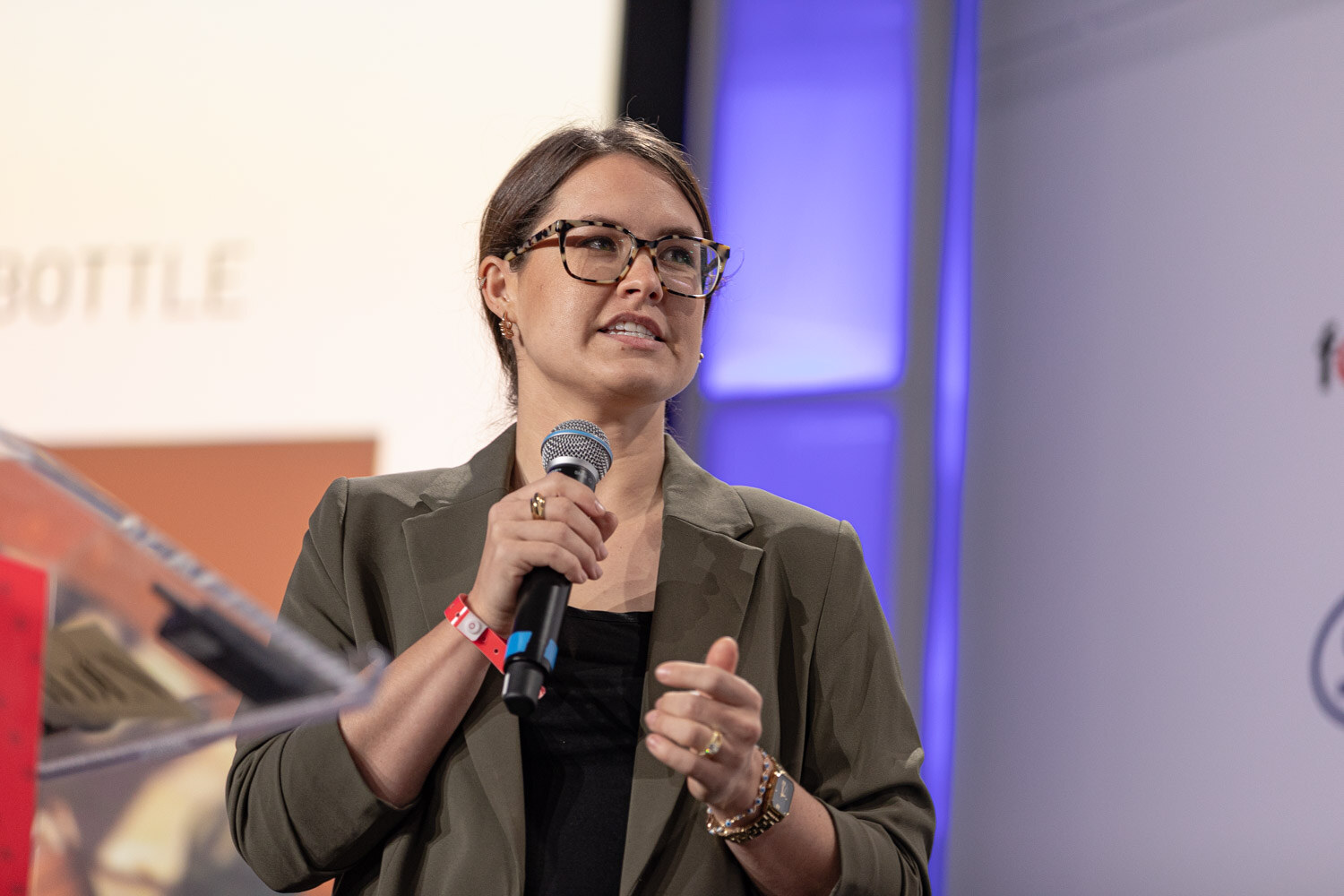 A woman in glasses and an olive blazer holds a microphone and gestures with her other hand beside a clear podium.