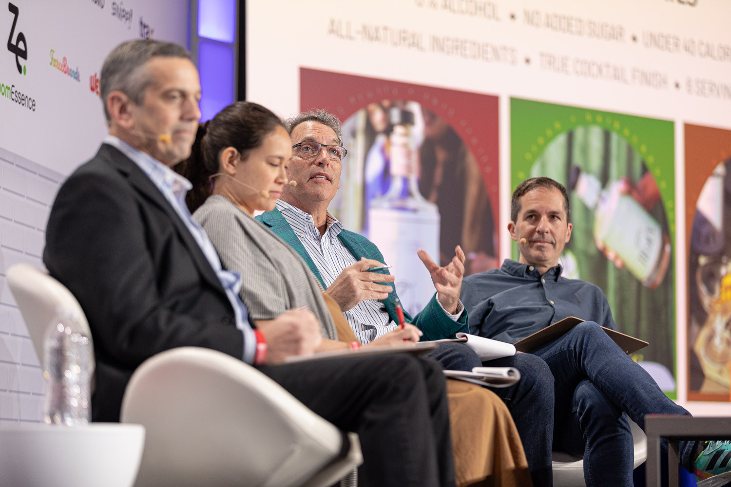 Four judges seated in white chairs, one in a teal blazer and one with a pen raised, in front of a screen showing bottles and all-natural ingredient claims.