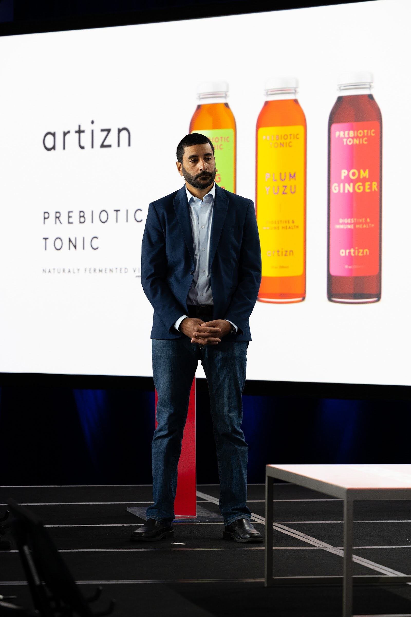 A man in a navy blazer and jeans stands on a dark stage in front of a white screen showing three artizn prebiotic tonic bottles.