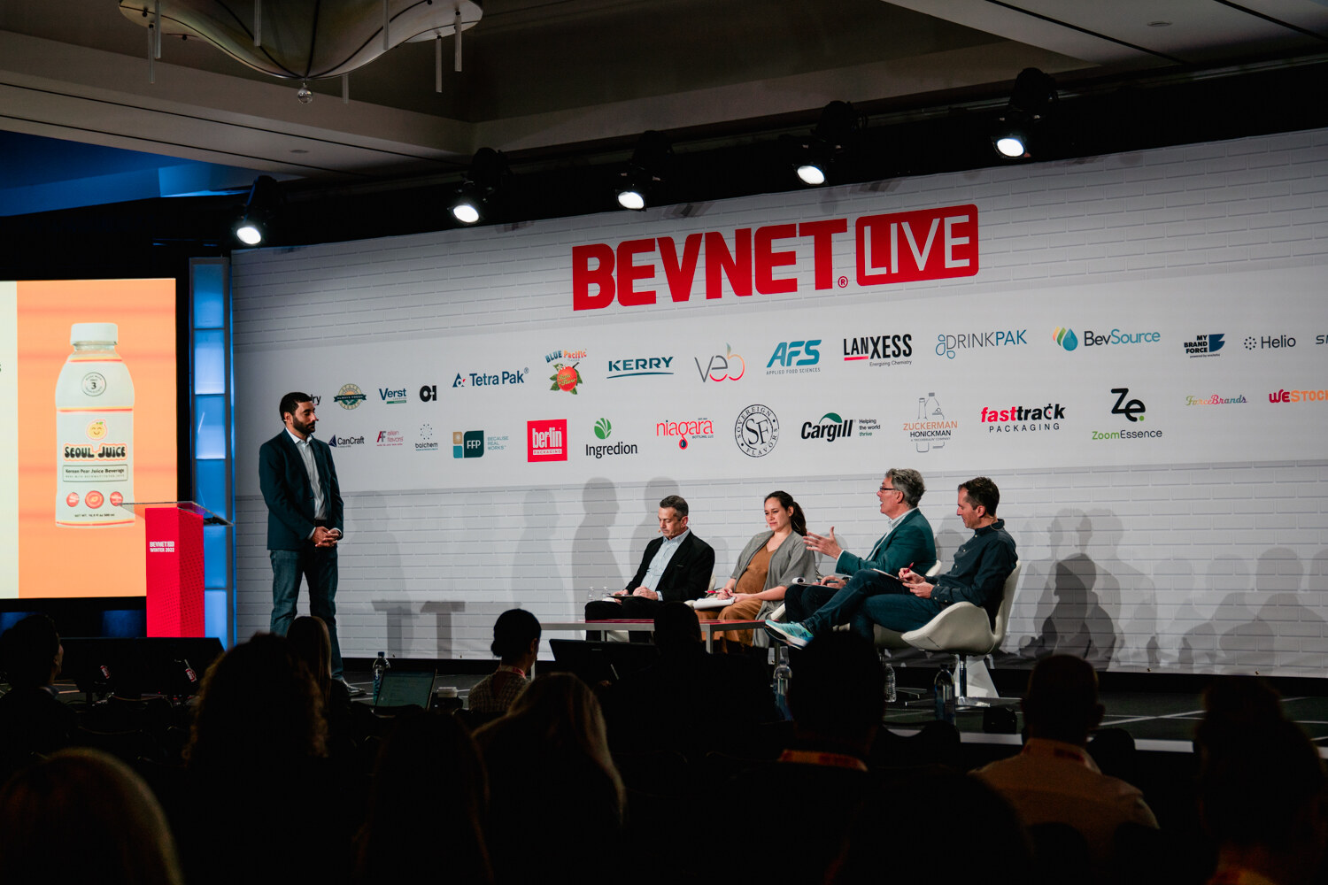 A wide shot of a stage showing a man standing at left, four judges seated in white chairs and a juice bottle on a screen under a BevNET Live sign.