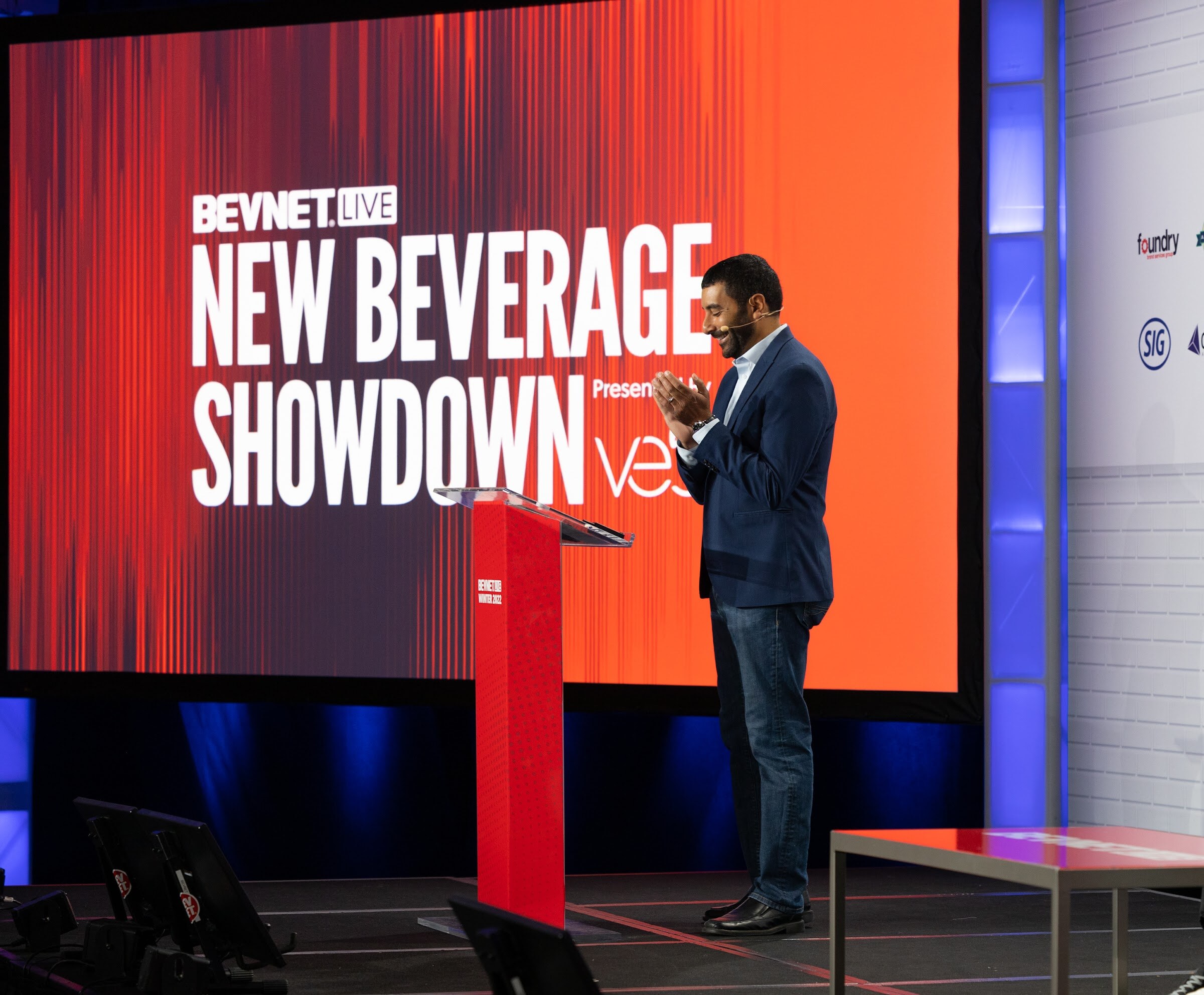 A man in a navy blazer claps beside a red podium in front of a large red New Beverage Showdown screen presented by Veb.