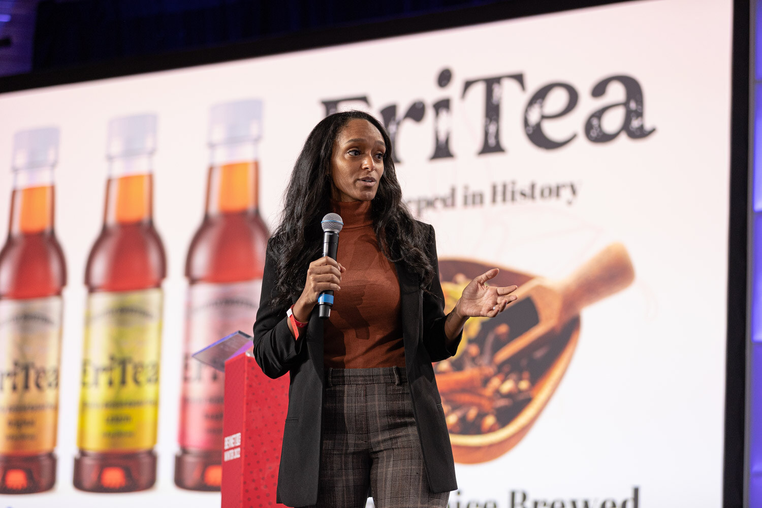 A woman with long dark hair in an orange top and black blazer holds a microphone in front of a screen reading EriTea, Steeped in History.