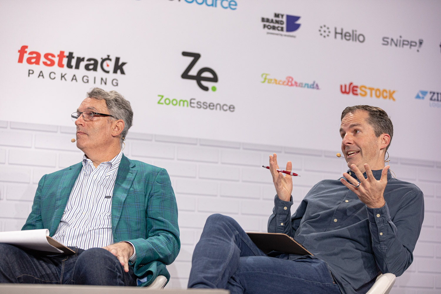 A man in a teal plaid blazer and a man in a navy shirt, both holding notes and pens, gesture as they talk in front of a sponsor wall.