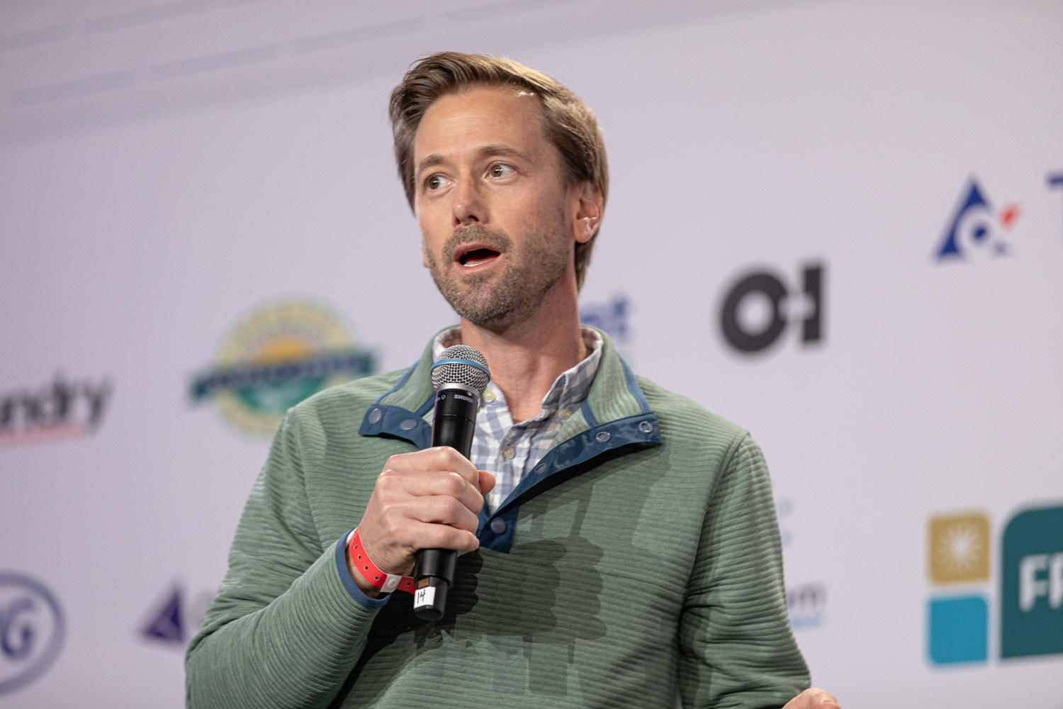 A bearded man in a green pullover holds a microphone with a red wristband in front of a blurred sponsor wall.