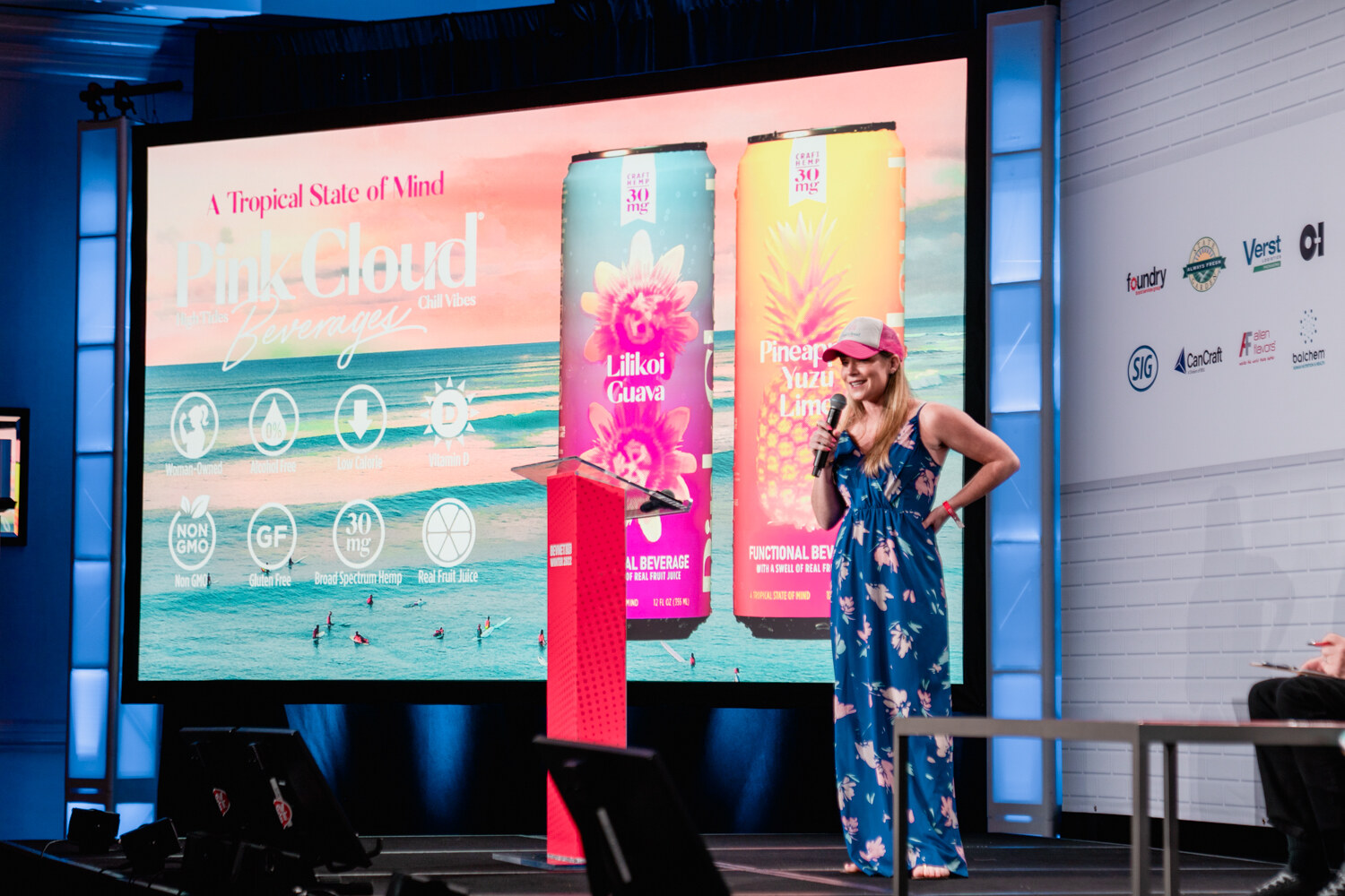 A woman in a floral dress stands at a podium on stage in front of a large screen reading Pink Cloud Beverages with two colorful cans.