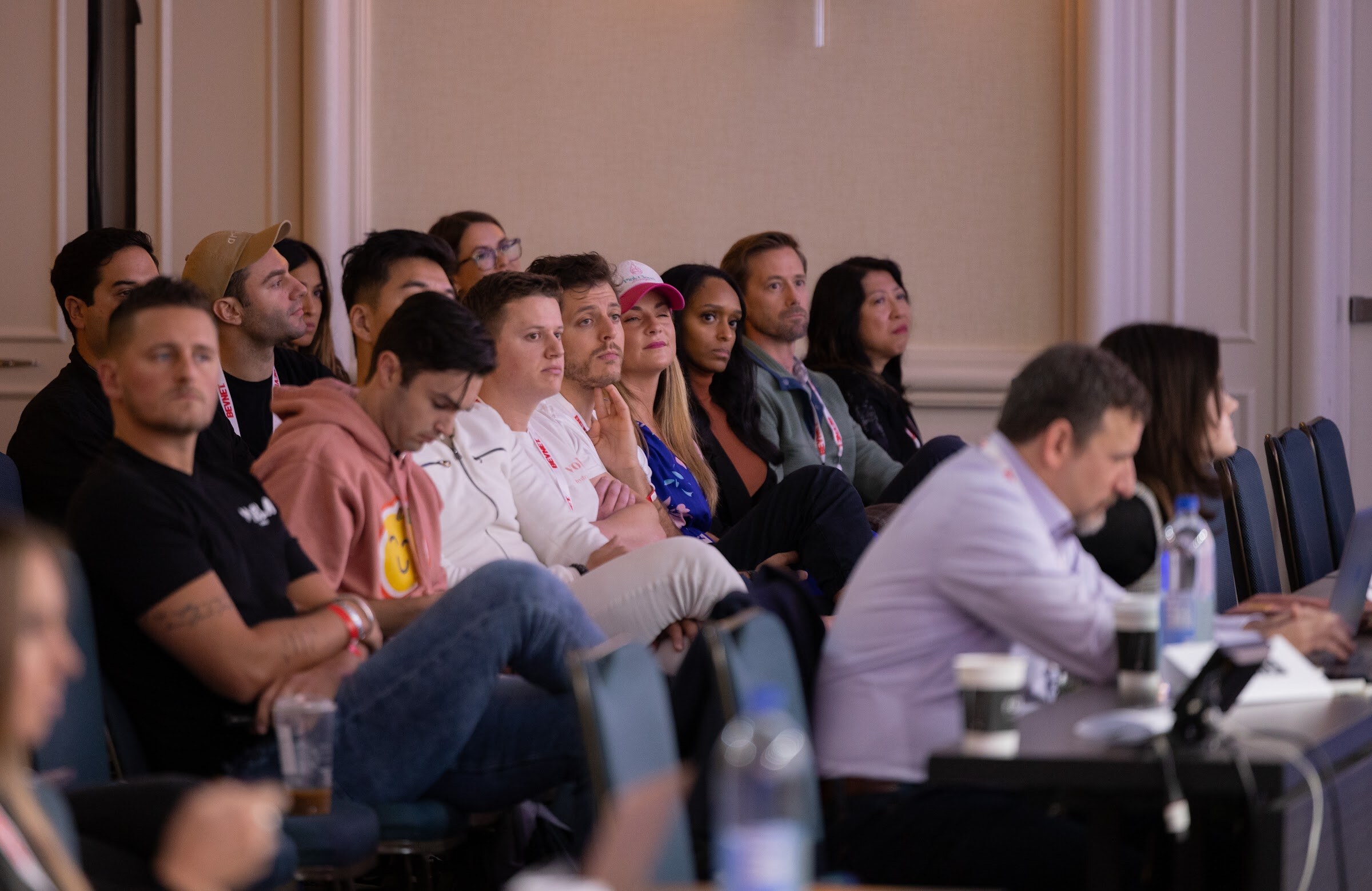 Seated attendees in a row watch the stage, with coffee cups and laptops on the table in front of them.