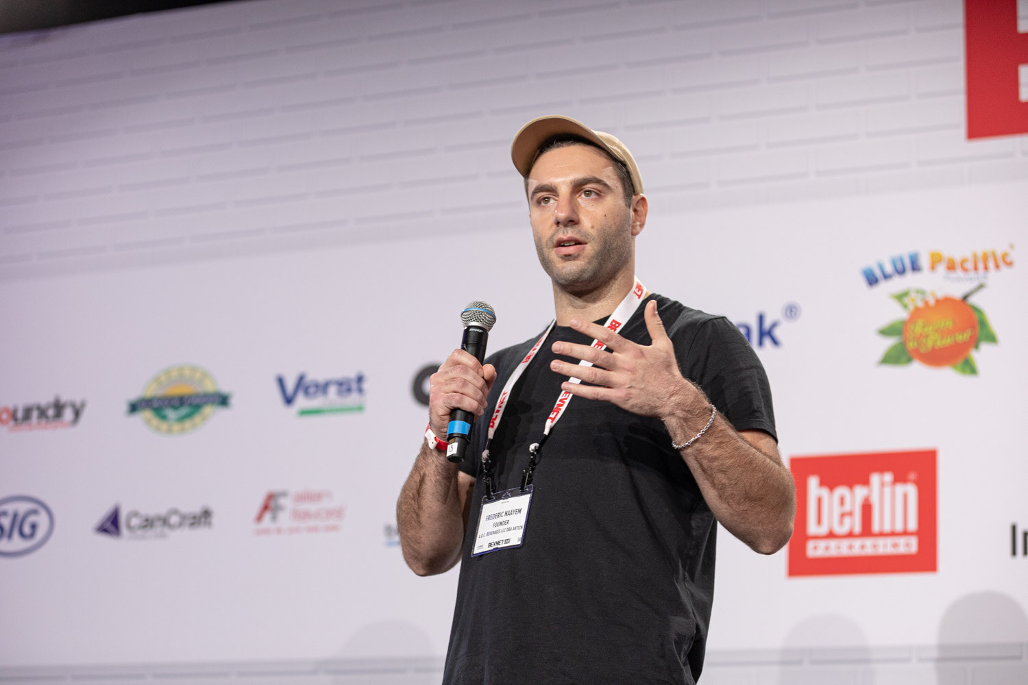 A man in a tan cap and black t-shirt with a BevNET lanyard holds a microphone and gestures with his other hand.