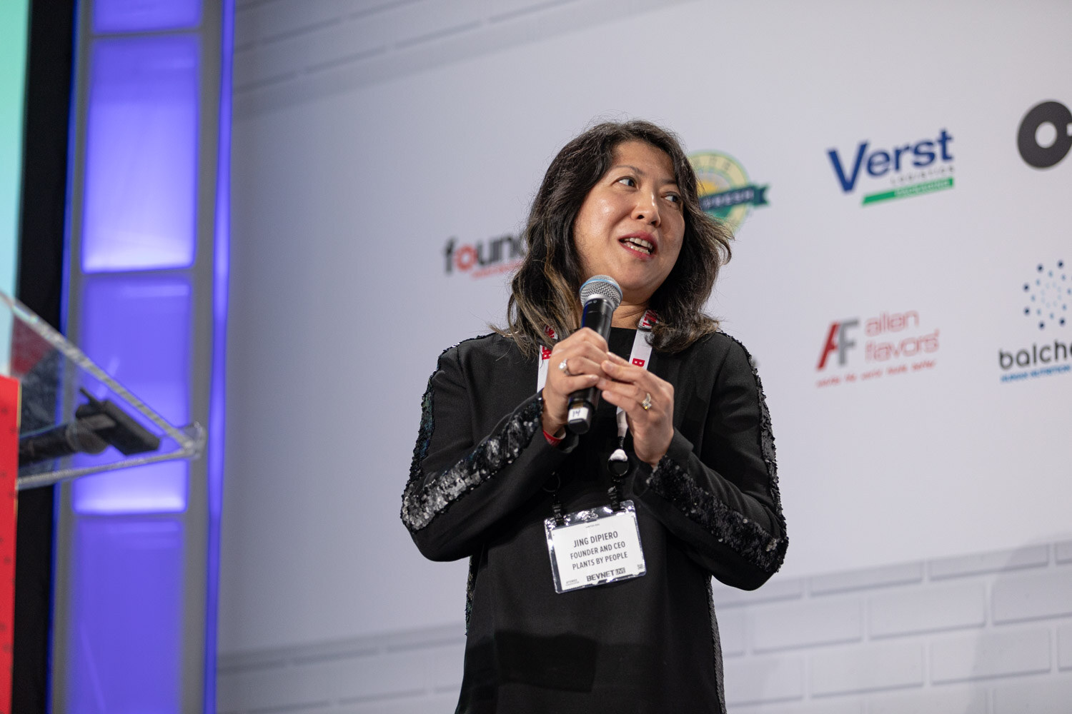 A woman in a black embroidered top with a badge holds a microphone beside a clear podium in front of a sponsor wall.