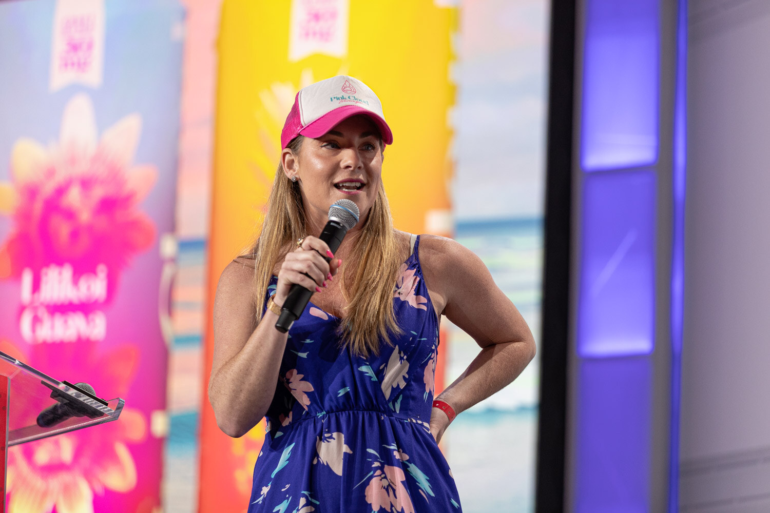 A woman with long blond hair in a pink and white cap and floral sundress holds a microphone and rests one hand on her hip in front of a colorful beach-themed screen.