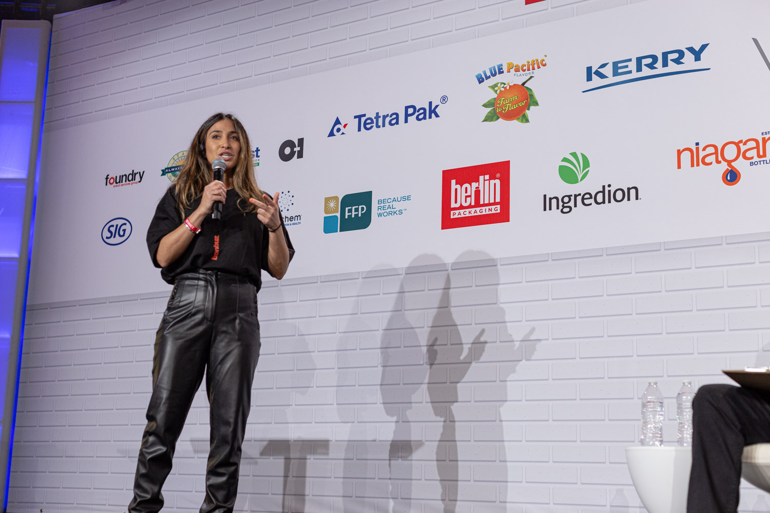 A woman in a black top and black leather pants holds a microphone on a stage in front of a white brick wall with sponsor logos.