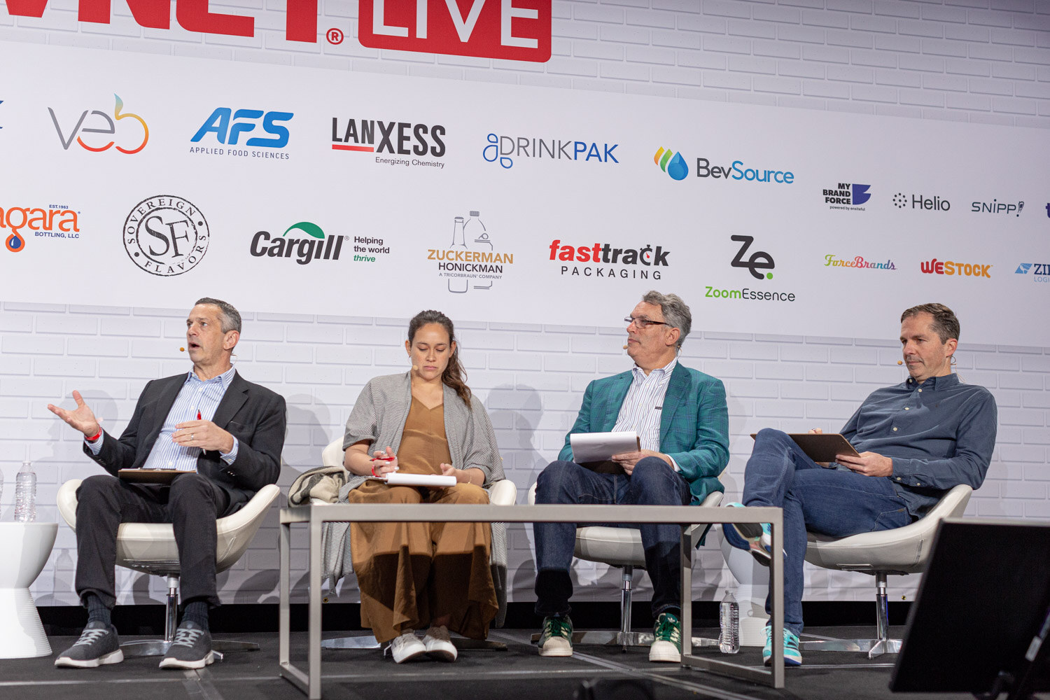 Four judges in white chairs behind a low table on a stage in front of a sponsor wall, each holding papers or a clipboard.
