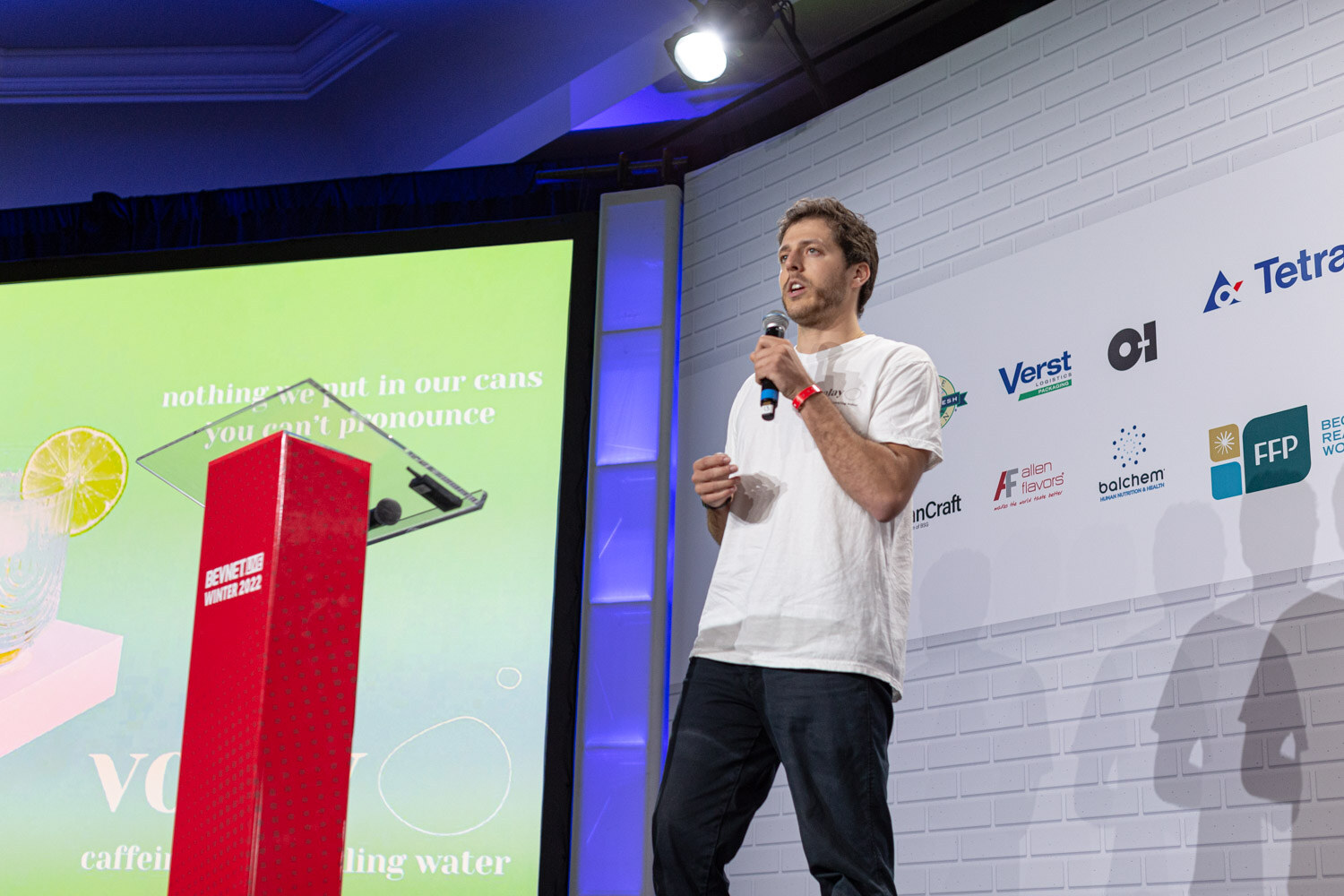 A man in a white t-shirt holds a microphone beside a red podium in front of a green screen reading nothing we put in our cans you can't pronounce.