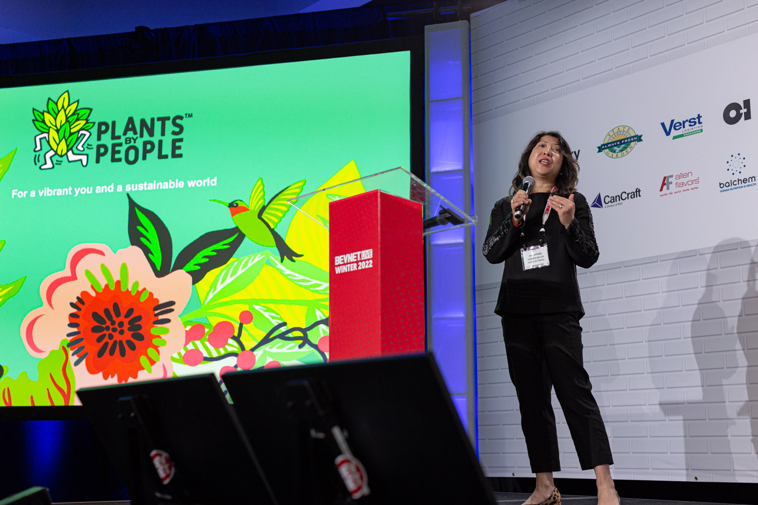 A woman in black stands in front of a bright green screen reading Plants by People, For a vibrant you and a sustainable world.