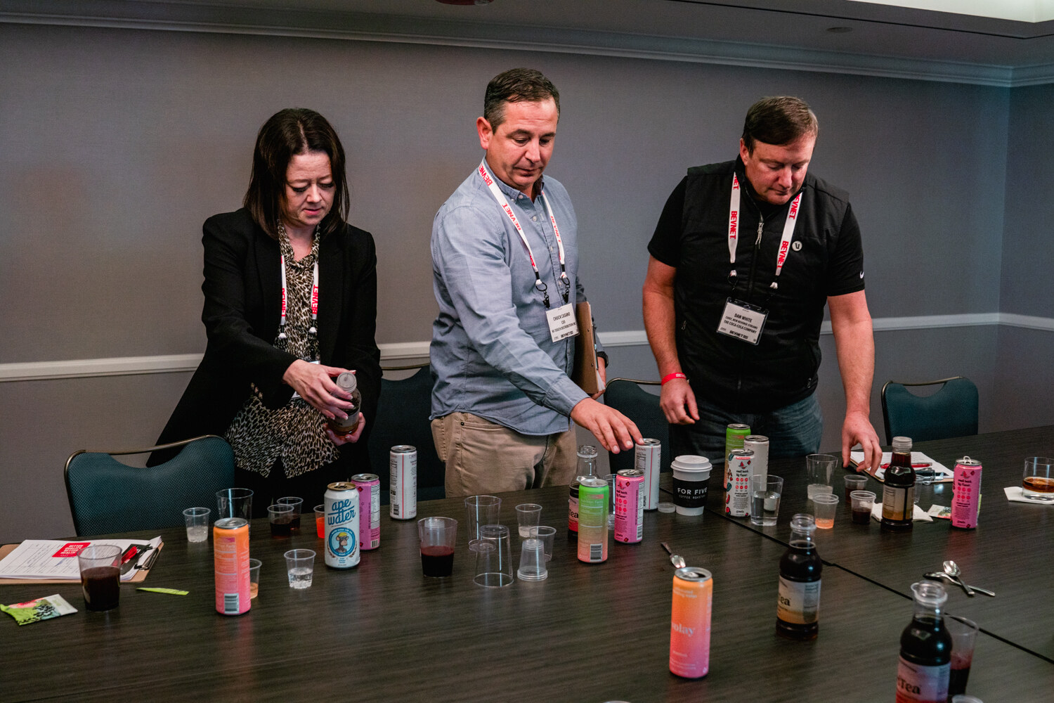 Three judges wearing BevNET lanyards stand at a long table covered with cans, bottles, plastic cups and scoring clipboards.