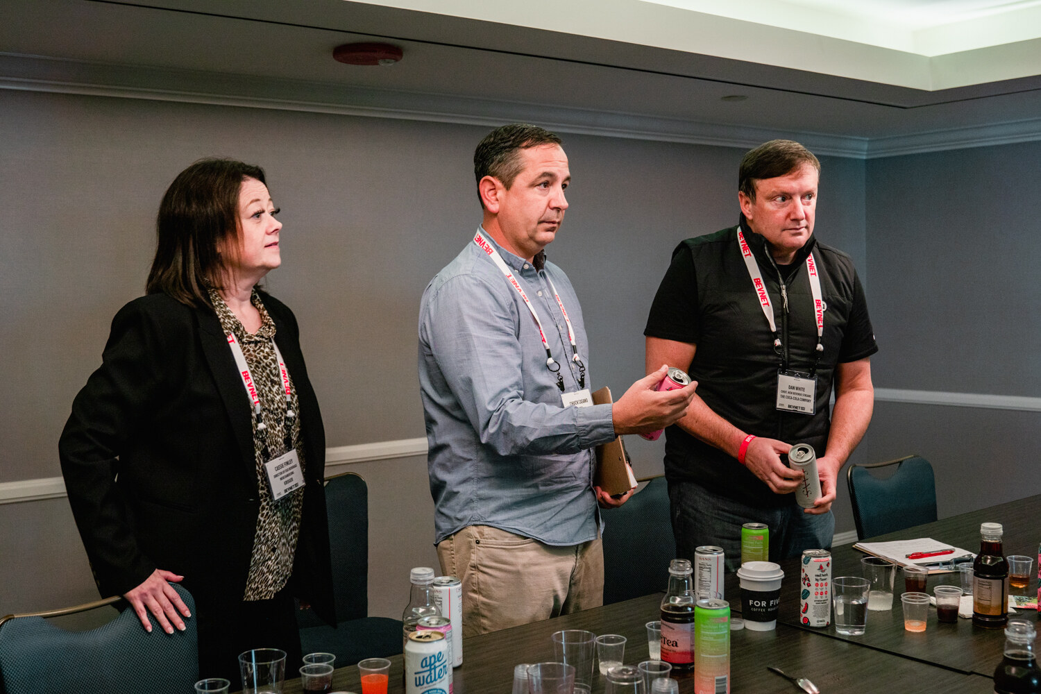 Three judges standing behind a table full of cans, bottles and cups, one holding a can and clipboard.