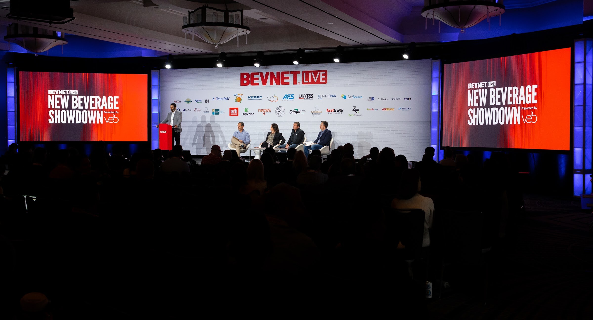 A dark ballroom with a stage in the center and two screens reading New Beverage Showdown presented by Veb flanking a BevNET Live sign.