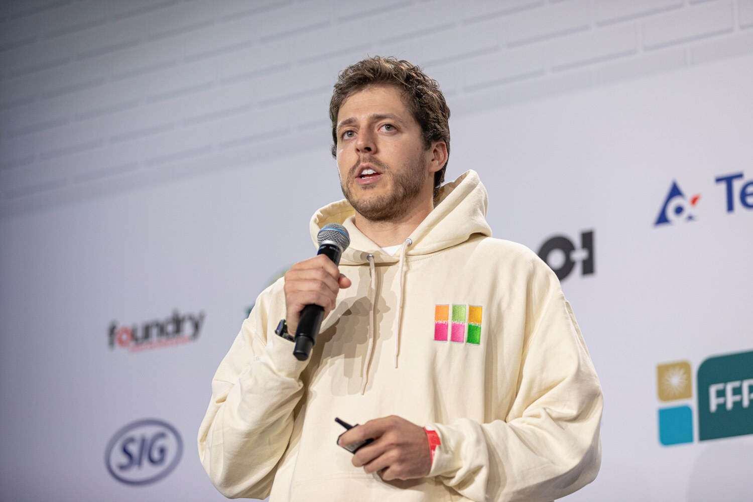 A bearded man in a cream hooded sweatshirt with a colorful square logo holds a microphone and clicker in front of a sponsor wall.