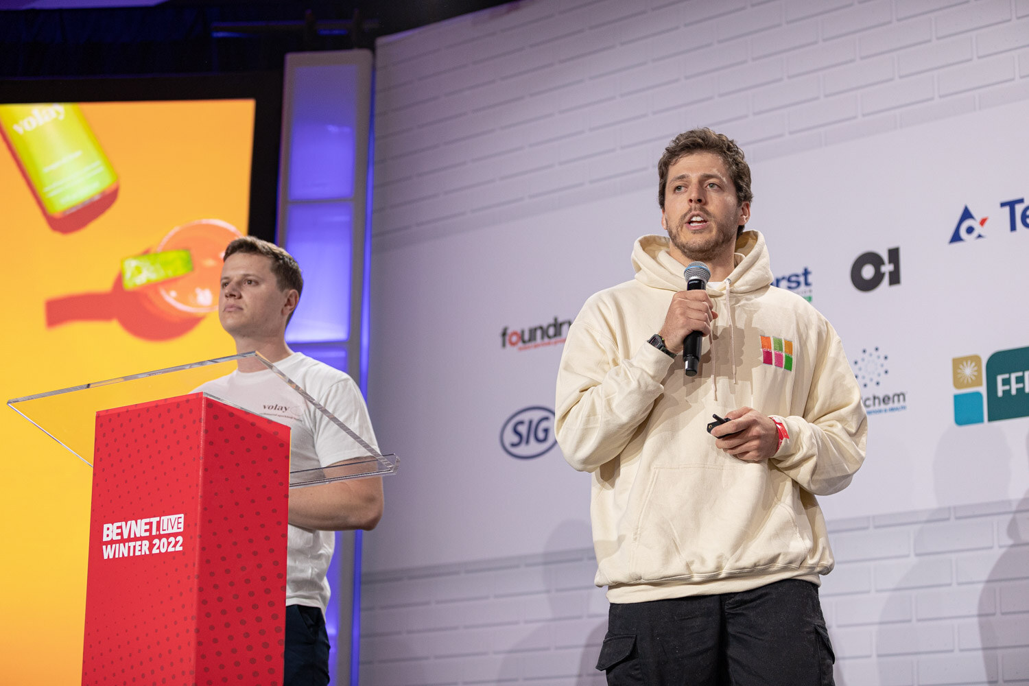 A man in a cream hoodie holds a microphone in the foreground while a man in a white t-shirt stands behind a red podium in front of an orange slide.