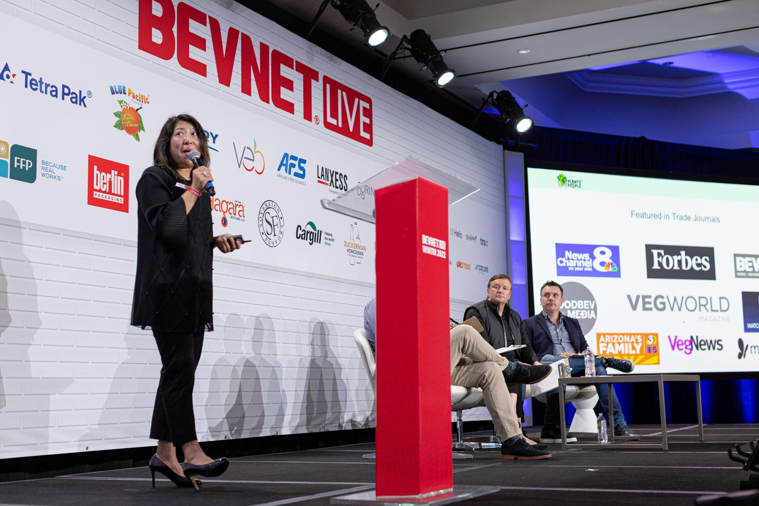 A woman in black stands beside a red podium and a large screen showing Featured in Trade Journals logos including Forbes, VegWorld and VegNews, with two judges seated behind her.