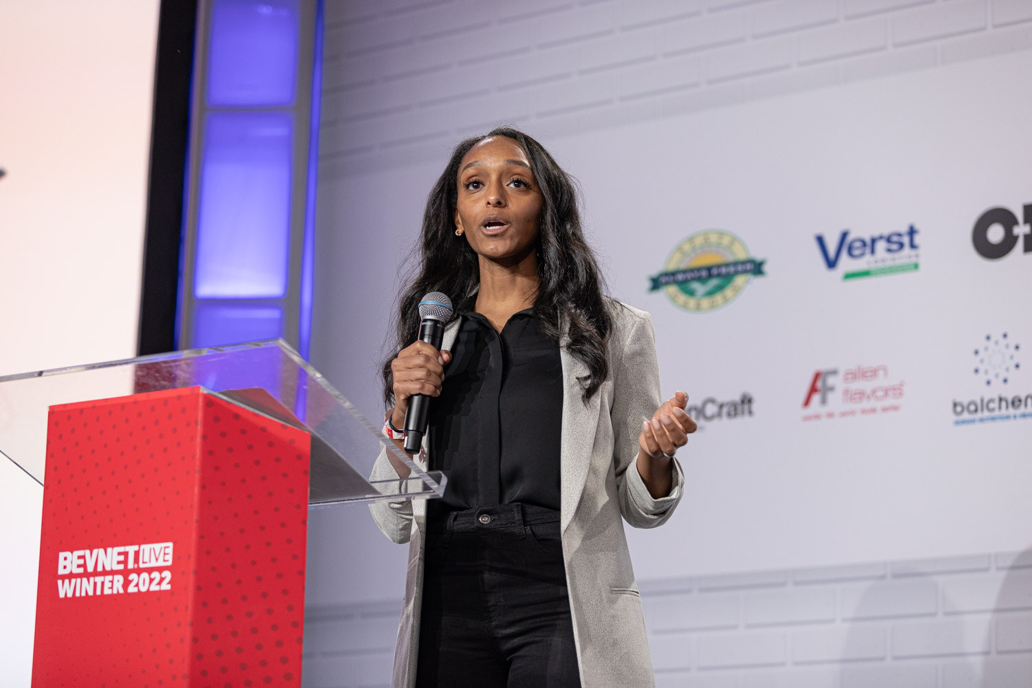 A woman with long dark hair in a gray blazer and black blouse holds a microphone beside a BevNET Live Winter 2022 podium.