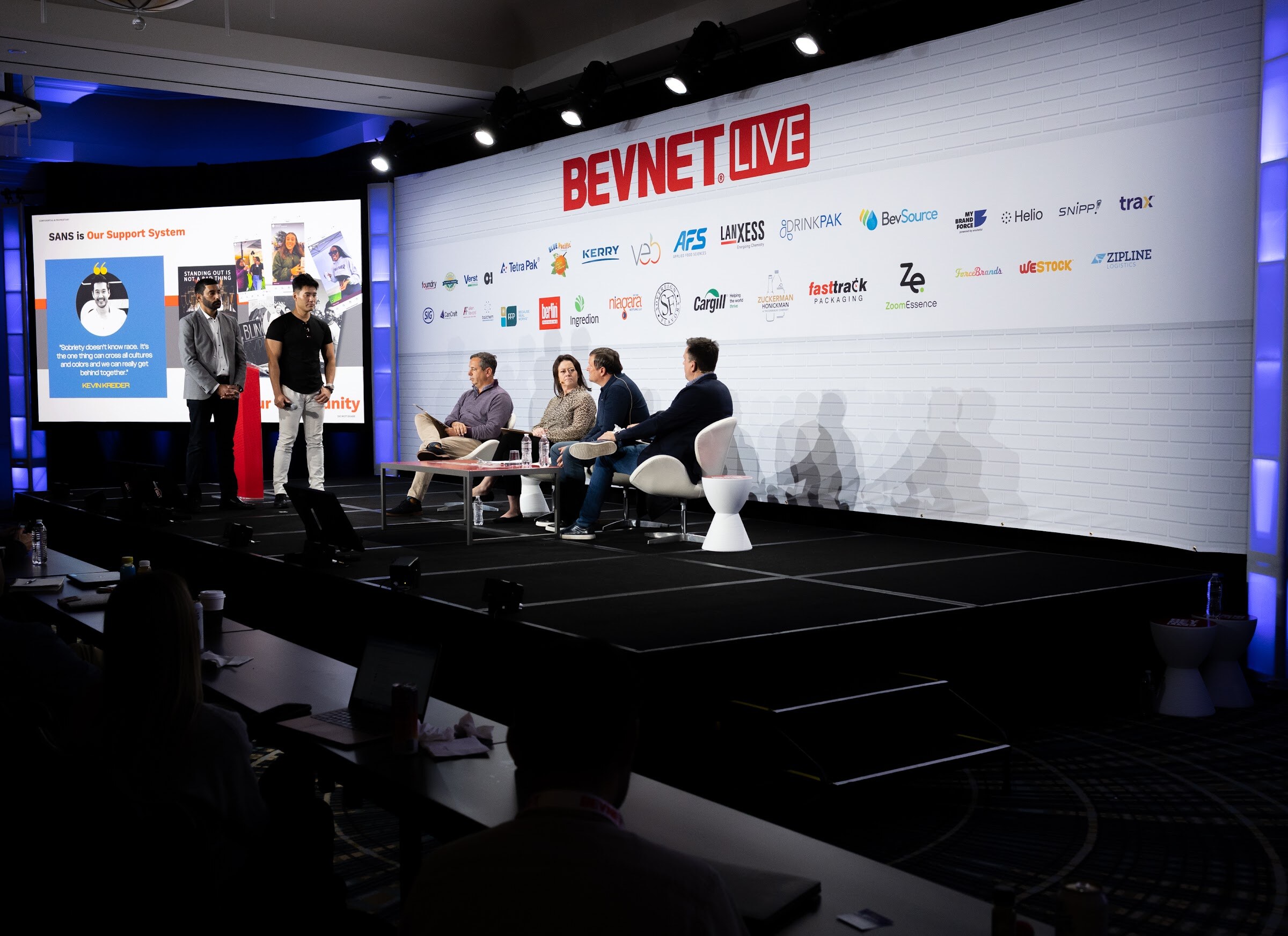A wide view of a stage with two men standing in front of a screen reading Sans is our support system, with four seated judges to the right.
