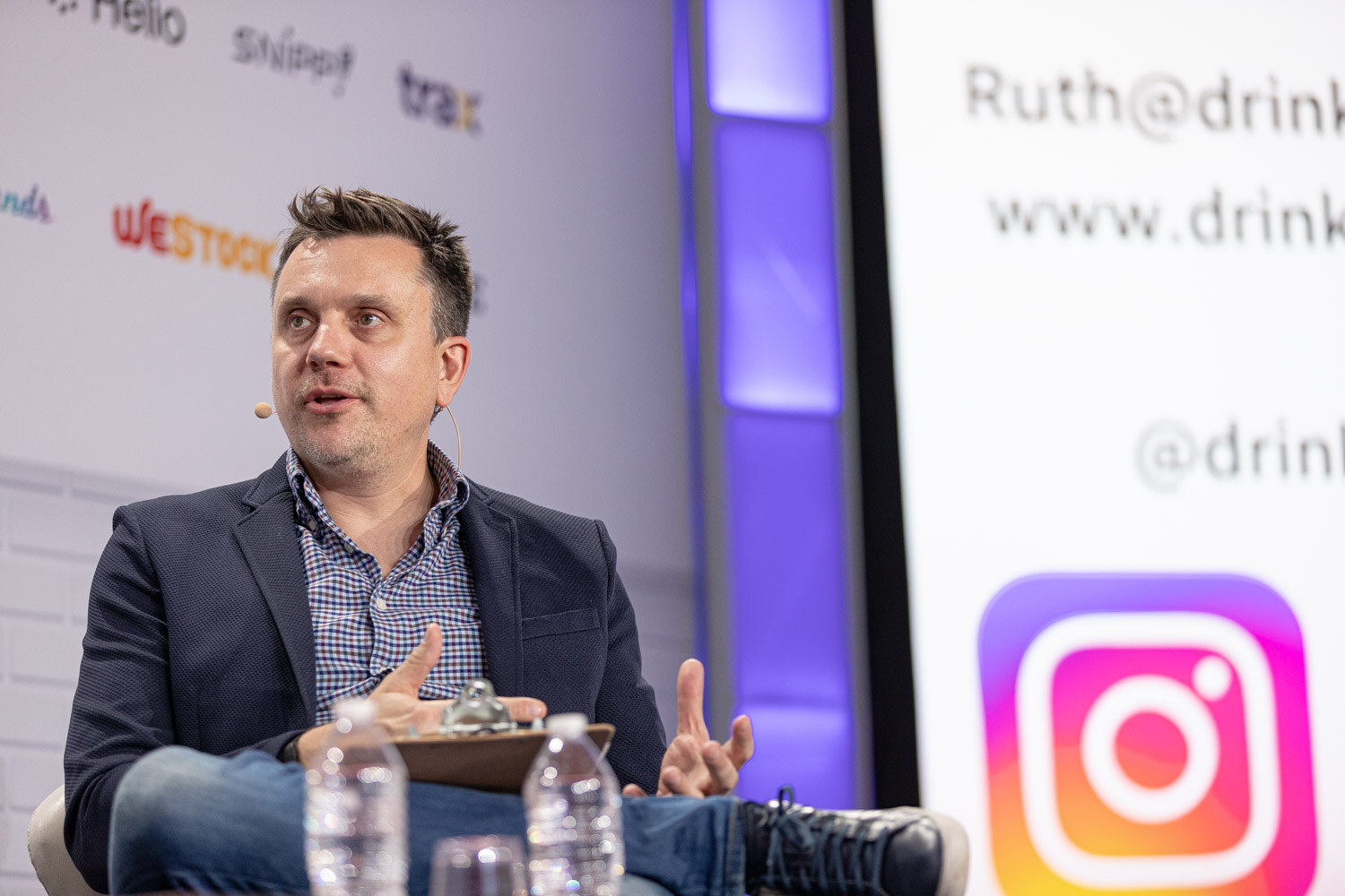 A man in a navy blazer and checked shirt sits with a clipboard and two water bottles next to a screen showing an Instagram icon and website address.