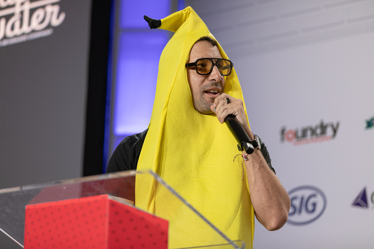 A man in a yellow banana costume and orange-tinted glasses holds a microphone behind a clear podium.