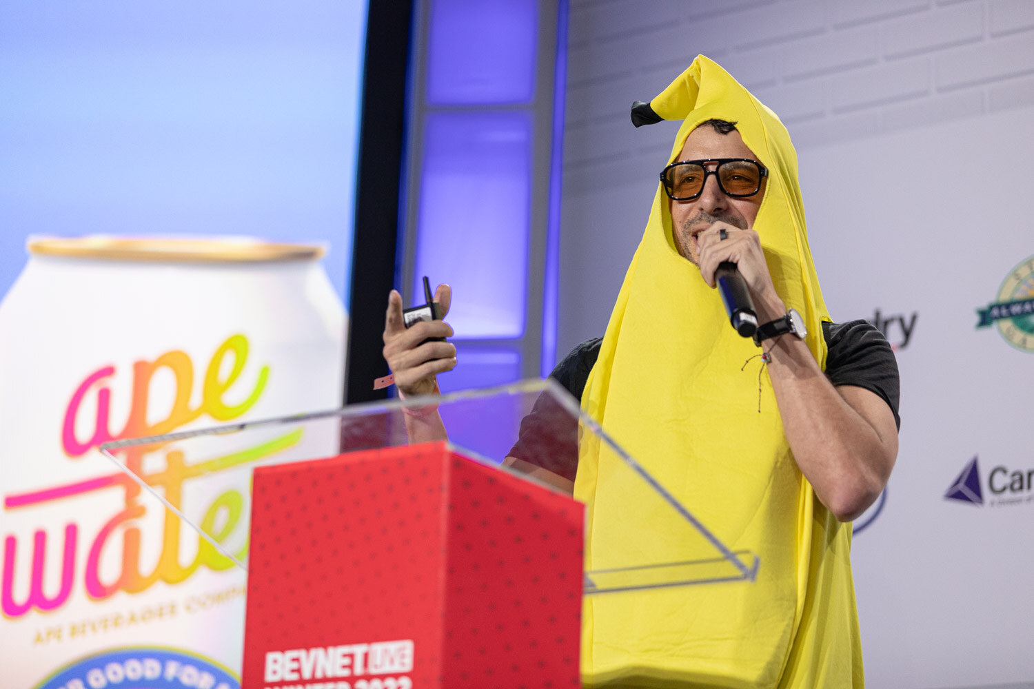 A man in a yellow banana costume and orange-tinted glasses holds a microphone and clicker at a red BevNET Live Winter 2022 podium in front of a screen showing an ape water can.