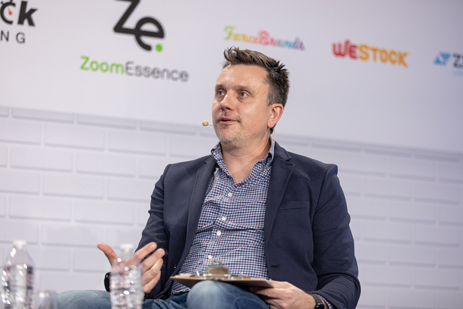 A man in a navy blazer with a headset microphone gestures while holding a clipboard in front of a ZoomEssence sponsor wall.