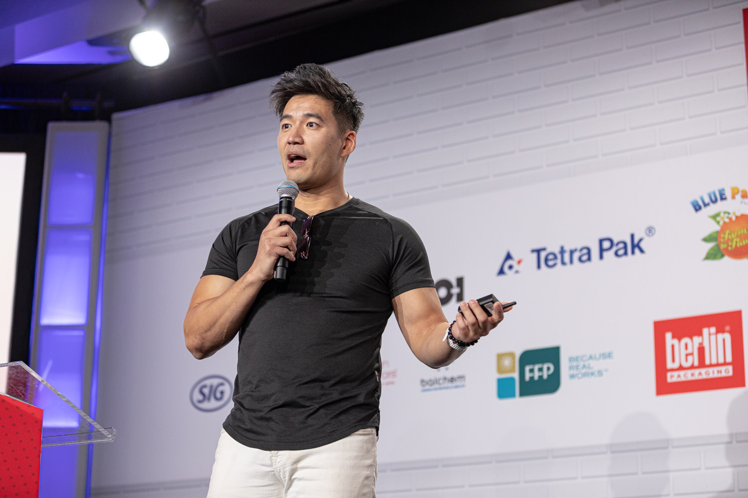 A man in a charcoal t-shirt and white pants holds a microphone in one hand and a phone in the other in front of a sponsor wall.
