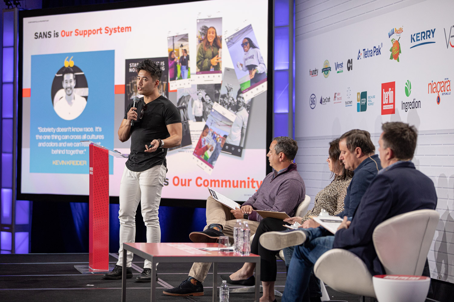A man in a black t-shirt and white pants stands at the left of the stage beside a slide reading Sans is Our Support System, with four judges seated to the right.