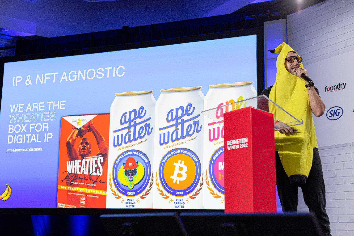 A person in a banana costume and orange glasses stands beside a screen reading IP & NFT agnostic with ape water cans and a Wheaties box.