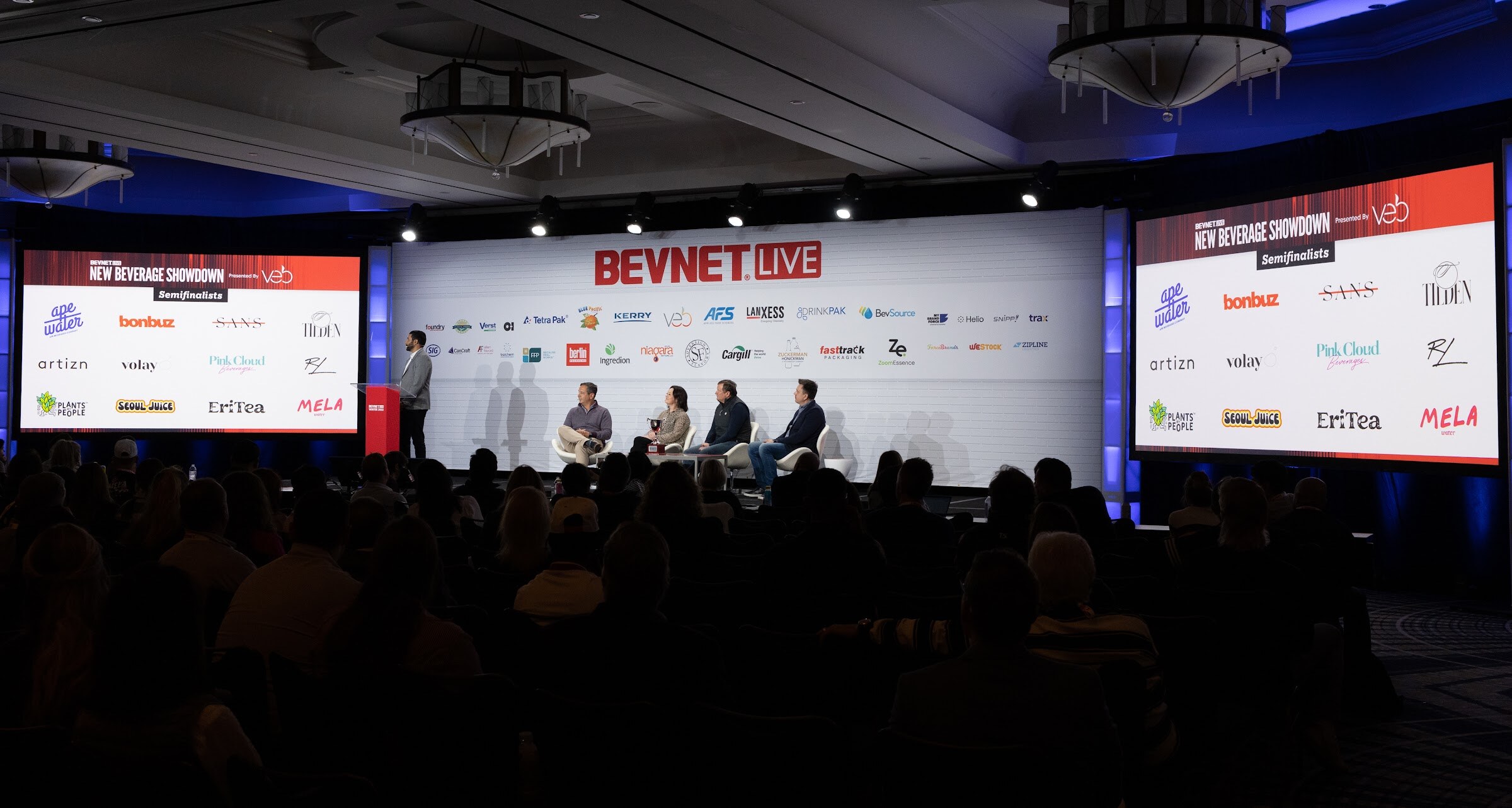 A wide view of the dark ballroom with two screens reading New Beverage Showdown presented by Veb listing the semifinalist brands, and four judges seated on stage.