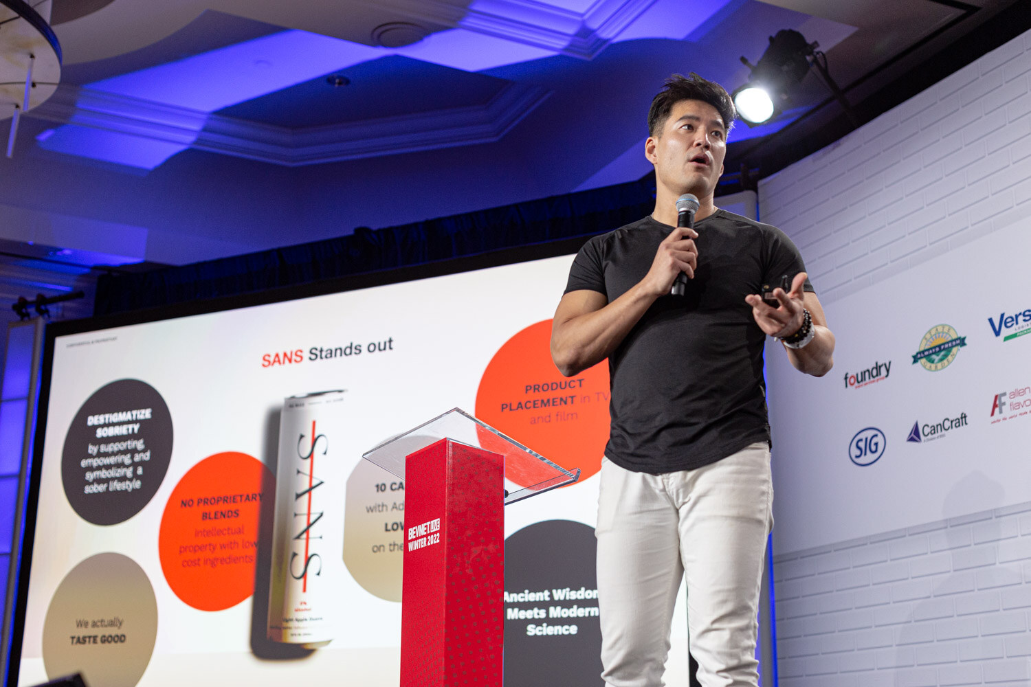 A man in a black t-shirt and white pants holds a microphone and clicker beside a red podium and a screen reading Sans Stands out.
